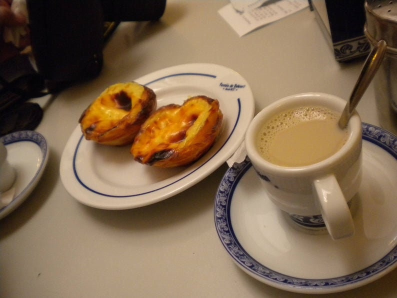 Puede incluir: Dos tartas de crema portuguesas doradas sobre un plato blanco con borde azul. Una taza de caf&eacute; con leche en una taza blanca con borde azul se encuentra sobre un platillo blanco con un patr&oacute;n azul y blanco.