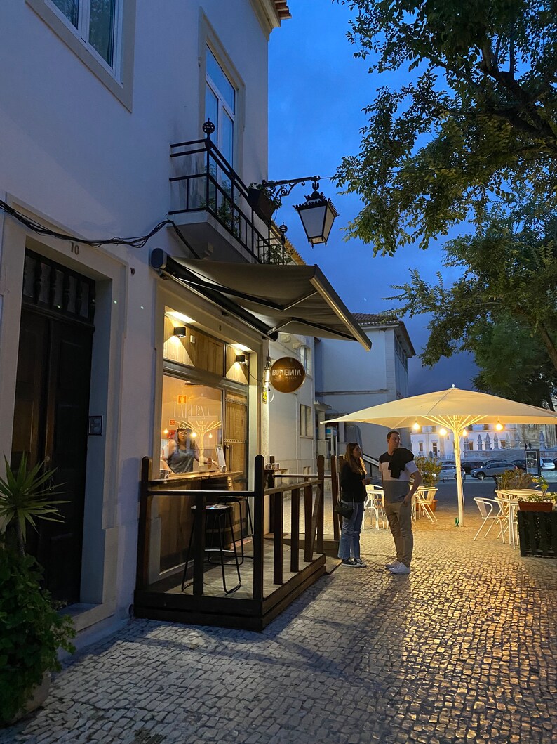May include: A small restaurant with a white awning and a sign that says "Bistr&ocirc;mia". There are two people standing outside the restaurant, and a white umbrella is set up on the patio.