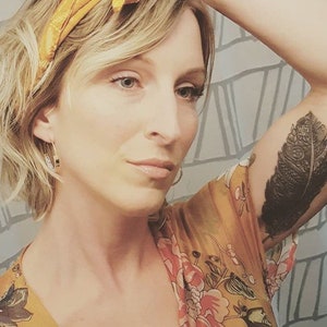 Peut inclure: Une femme aux cheveux blonds courts portant un bandeau jaune et une robe &agrave; fleurs. Elle a un tatouage de plume noire sur le haut du bras.