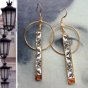 May include: A pair of gold hoop earrings with a dangling rectangular charm. The charms are white with a black floral pattern.