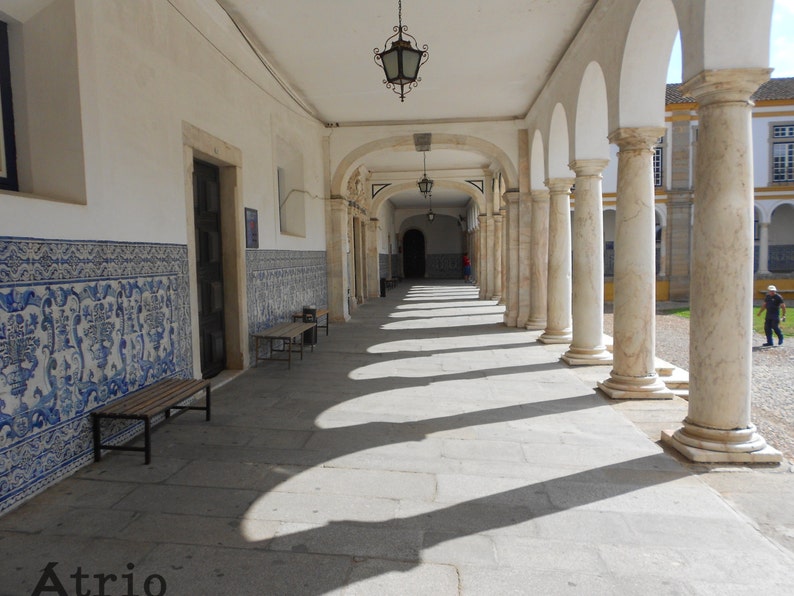 Peut inclure: Un b&acirc;timent blanc avec une longue all&eacute;e couverte bord&eacute;e de colonnes blanches. L'all&eacute;e est carrel&eacute;e et a un mur carrel&eacute; bleu et blanc sur le c&ocirc;t&eacute; gauche. Le texte "Atrio" est visible sur le sol.