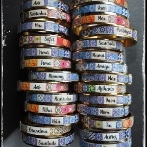May include: A stack of colorful bangle bracelets with Portuguese tile designs and gold accents. Each bracelet features a different word in Portuguese, such as "Vovó", "Mãe", "Amiga", and "Prima".