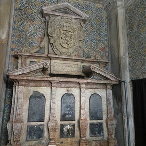 May include: A stone fountain with a carved coat of arms above it. The fountain has three arched openings and a spout in the center. The inscription on the fountain reads "DE DOMPE VIRTVTEM MA NIBVS MEIS".