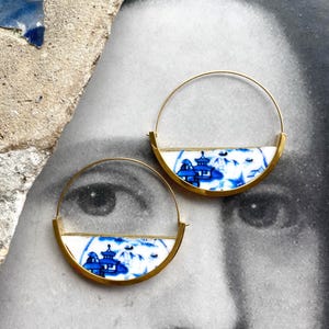 May include: A pair of gold-tone hoop earrings. Each earring features a half-circle of white porcelain with a blue landscape design. The earrings are set against a black and white background.
