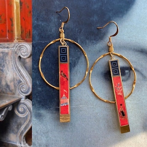 May include: A pair of gold hoop earrings with a red and black rectangular pendant. Each pendant features a different design with a bird, a building, and flowers.