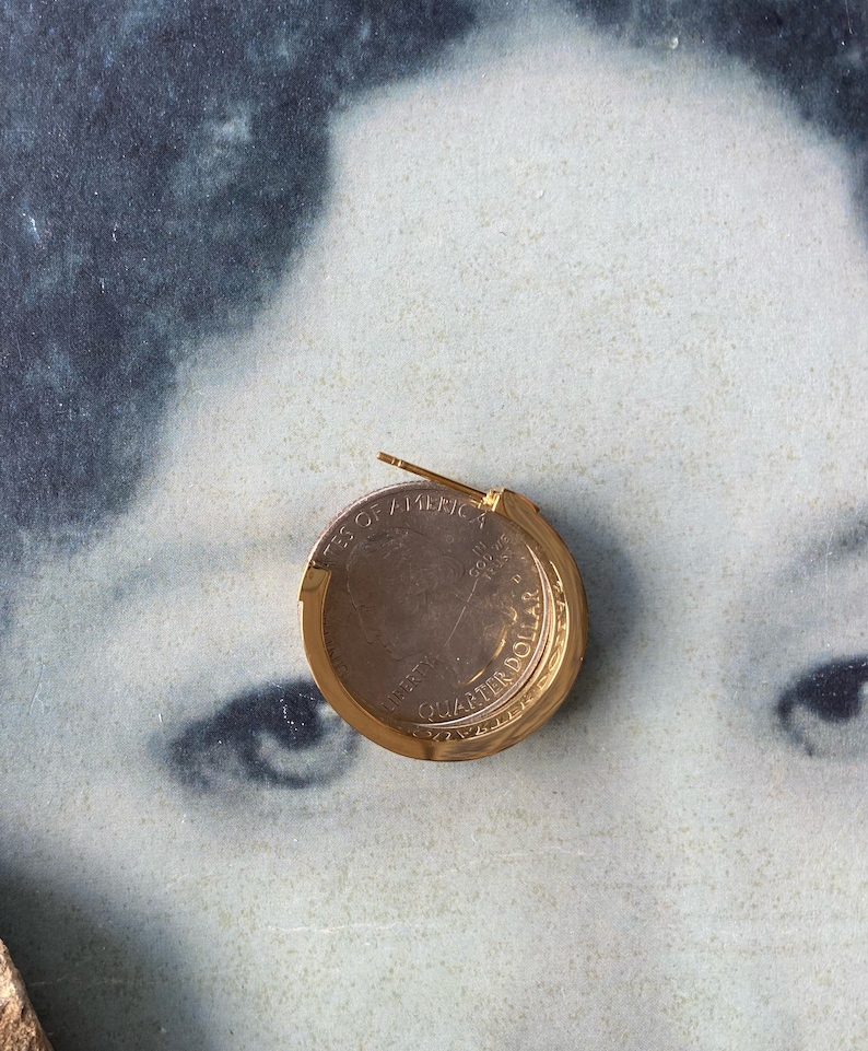 Op de afbeelding: Een gouden oorknop in de vorm van een kwart dollar munt met de inscriptie "United States of America" en "Quarter Dollar" zichtbaar.