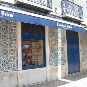 Op de afbeelding: Een winkelgevel met een blauwe luifel waarop "Pasteis de Belem - desde 1937" in het Portugees staat. De gevel is versierd met blauwe en witte tegels en heeft een grote etalage.