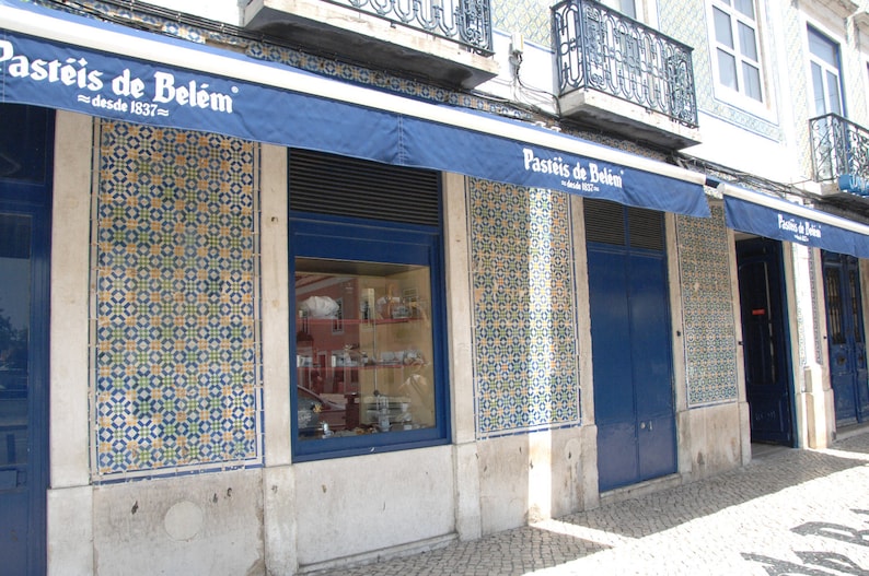 Op de afbeelding: Een winkelgevel met een blauwe luifel waarop "Pasteis de Belem - desde 1937" in het Portugees staat. De gevel is versierd met blauwe en witte tegels en heeft een grote etalage.