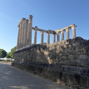 May include: The ancient Roman temple ruins in Evora, Portugal, feature a series of tall, weathered stone columns with ornate Corinthian capitals standing atop a high, rectangular stone podium constructed from large, rough-hewn granite blocks.