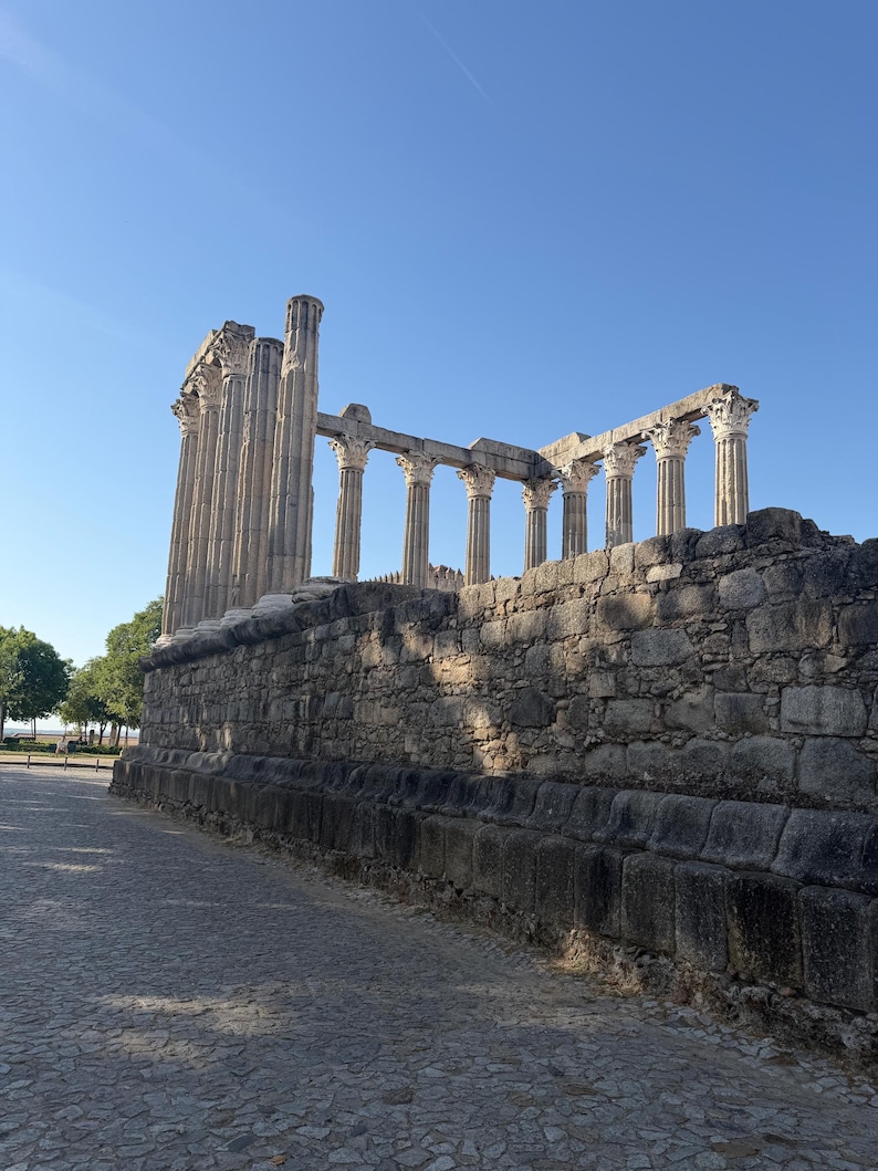 May include: The ancient Roman temple ruins in Evora, Portugal, feature a series of tall, weathered stone columns with ornate Corinthian capitals standing atop a high, rectangular stone podium constructed from large, rough-hewn granite blocks.