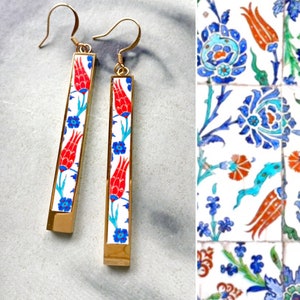 May include: A pair of gold earrings with a white background and red and blue floral designs. The earrings are long and rectangular in shape.