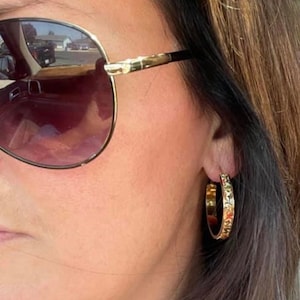 May include: A woman wearing gold hoop earrings with a textured design. She is wearing sunglasses and has long brown hair.