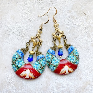 May include: A pair of ornate earrings with a gold-tone metal design. The earrings feature a crescent shape with blue and green patterned tiles, red accents, and a white bird design. Blue teardrop beads dangle from the bottom.