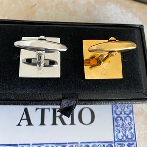 May include: A pair of cufflinks in a black box. One cufflink is silver and the other is gold. Both cufflinks have the word "ATRIO" engraved on them.