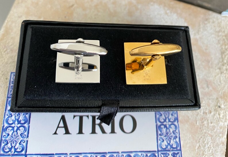 May include: A pair of cufflinks in a black box. One cufflink is silver and the other is gold. Both cufflinks have the word "ATRIO" engraved on them.
