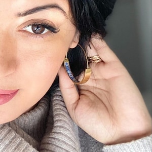 May include: Close-up of a person's face wearing gold hoop earrings with a blue and white patterned design. The earrings are large and have a wide band.