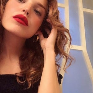 May include: A woman with long, curly, auburn hair wearing a black off-the-shoulder top. She is looking at the camera and has red lipstick on.