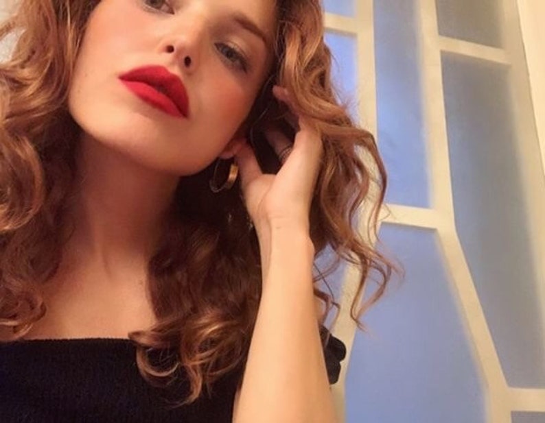 May include: A woman with long, curly, auburn hair wearing a black off-the-shoulder top. She is looking at the camera and has red lipstick on.