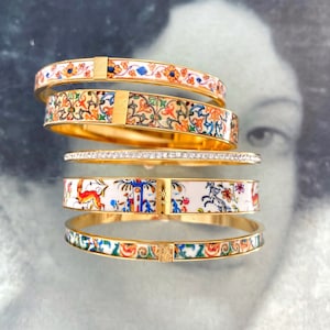 May include: A collection of gold-toned bangles with intricate designs. Some feature colorful floral patterns, others have a white background with illustrations of animals and buildings. One bangle is adorned with sparkling crystals.