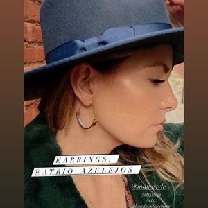 May include: A woman wearing a blue fedora hat and a green fluffy jacket. She is wearing gold hoop earrings with a blue and white design. The text "EARRINGS: @ATRIO AZULEJOS" is on the image. The text "@makastyle" is on the image. The text "#etsyshop #etsy #handmadejewelry #atrio #nycblogger #fallfashion #etsyjewelry #jewelrylover" is on the image.