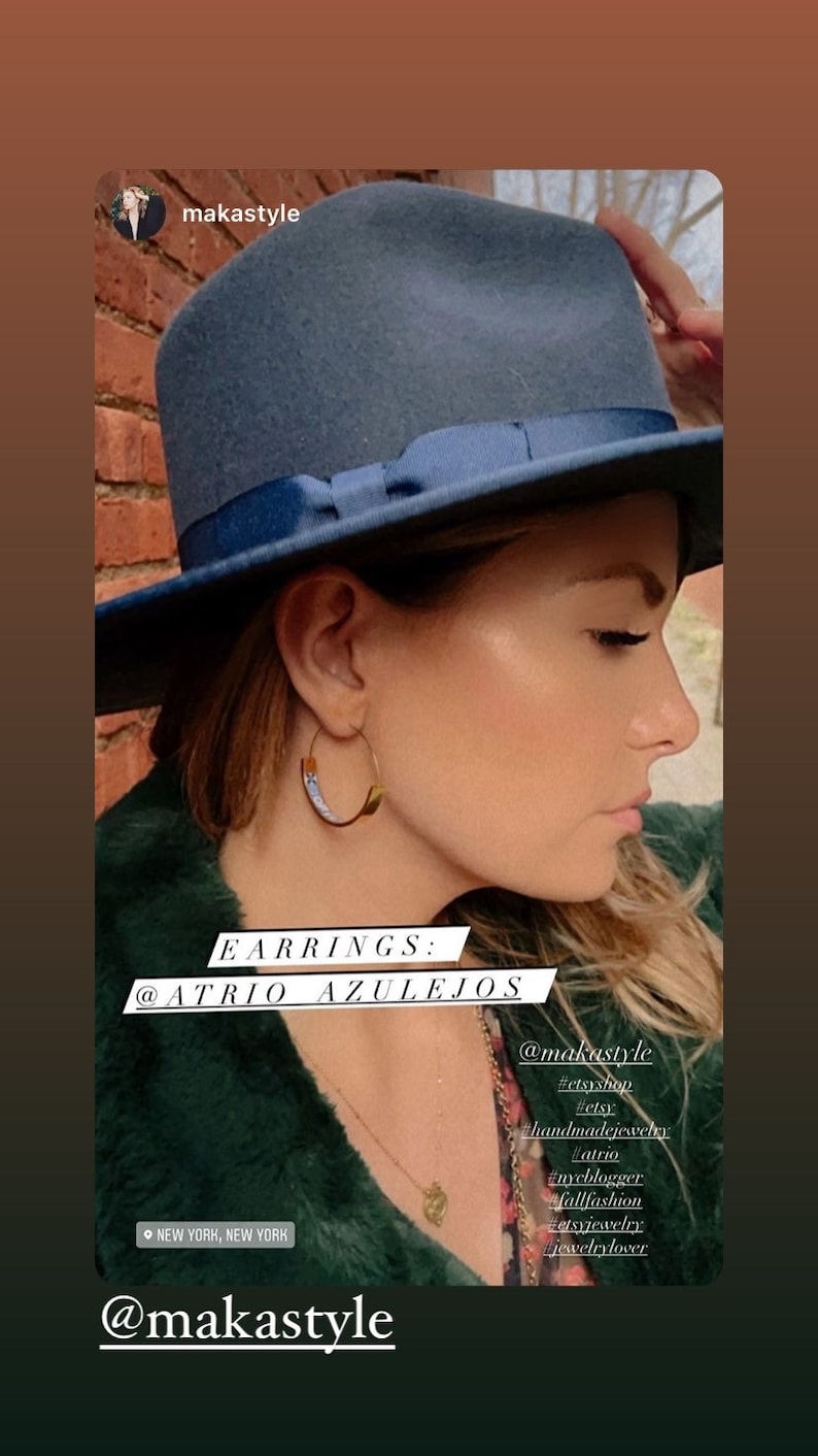 May include: A woman wearing a blue fedora hat and a green fluffy jacket. She is wearing gold hoop earrings with a blue and white design. The text "EARRINGS: @ATRIO AZULEJOS" is on the image. The text "@makastyle" is on the image. The text "#etsyshop #etsy #handmadejewelry #atrio #nycblogger #fallfashion #etsyjewelry #jewelrylover" is on the image.