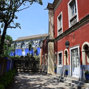 May include: A red building with white trim and blue and white tiled accents. The building has arched windows and a white door. The building is surrounded by a courtyard with a fountain and trees.