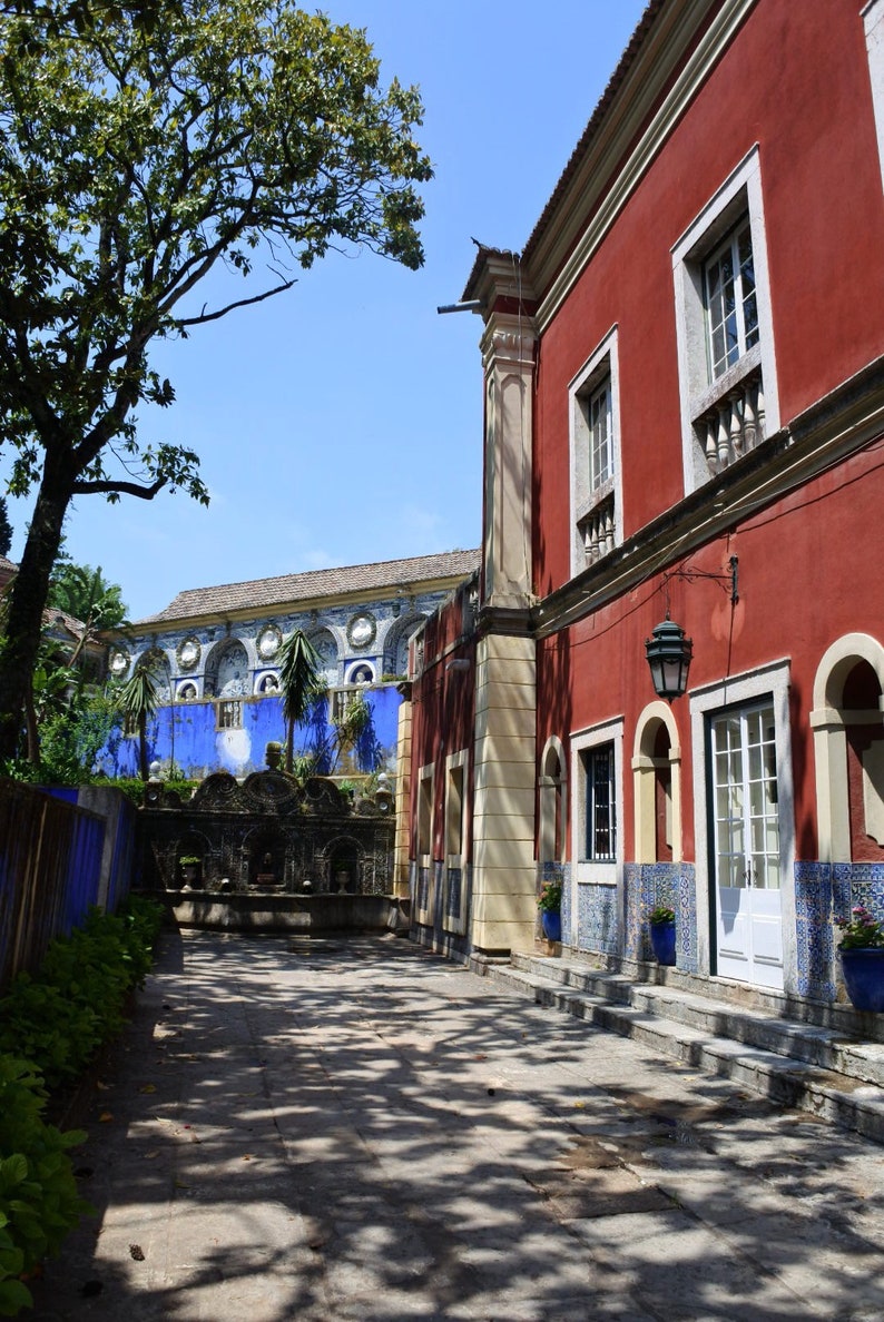 May include: A red building with white trim and blue and white tiled accents. The building has arched windows and a white door. The building is surrounded by a courtyard with a fountain and trees.