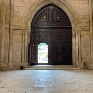 May include: A large, wooden door with intricate carvings and metal accents opens to reveal a view of a courtyard. The door is set within a stone archway, and the floor is made of large, square tiles.