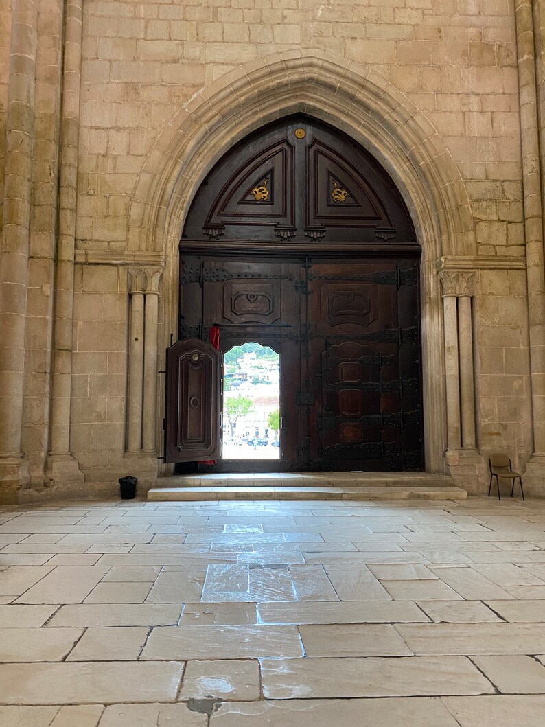 May include: A large, wooden door with intricate carvings and metal accents opens to reveal a view of a courtyard. The door is set within a stone archway, and the floor is made of large, square tiles.