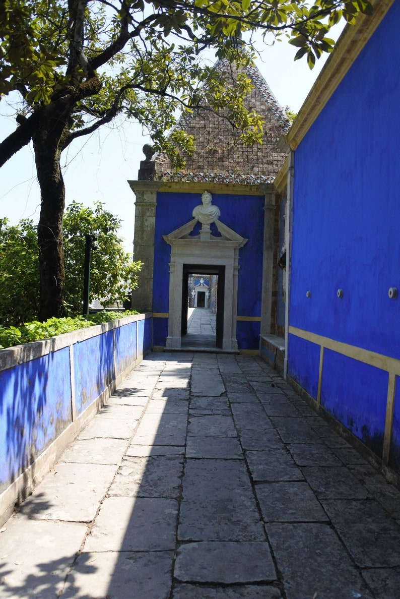 May include: A stone walkway leads to a doorway with a blue and white archway. The walls are painted a vibrant blue with yellow trim. The walkway is shaded by a tree.