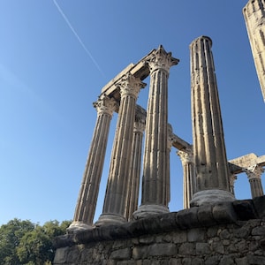 May include: Ancient stone ruins featuring tall, fluted columns with ornate Corinthian capitals standing atop a weathered stone base against a clear blue sky. The structure displays architectural remnants of a classical temple with vertical grooves.