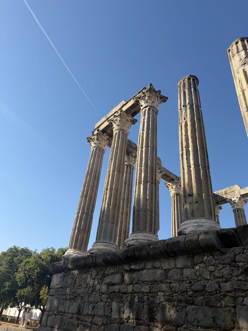 May include: Ancient stone ruins featuring tall, fluted columns with ornate Corinthian capitals standing atop a weathered stone base against a clear blue sky. The structure displays architectural remnants of a classical temple with vertical grooves.