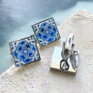 May include: A pair of cufflinks with a blue and white tile design. The cufflinks are made of silver and have a square shape.