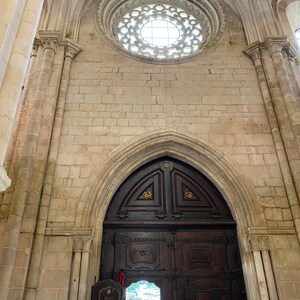 May include: A large, ornate wooden door with intricate carvings and metal accents. The door is set within a stone archway and leads into a building with a stone ceiling and a circular window.