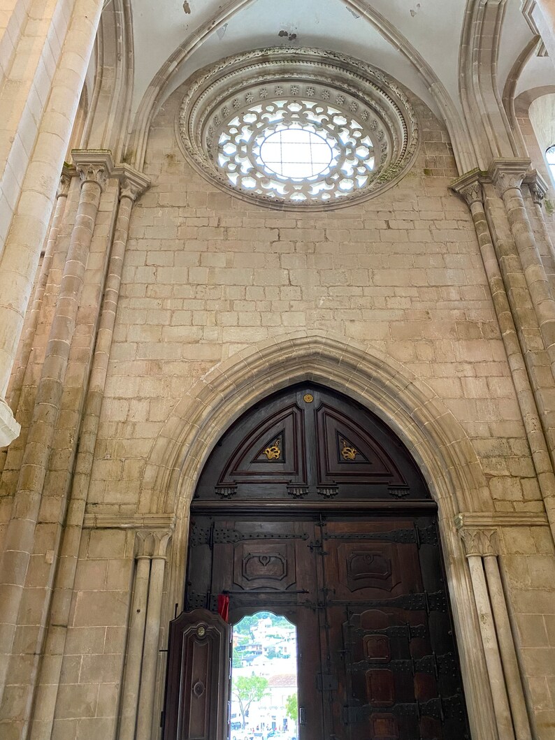 May include: A large, ornate wooden door with intricate carvings and metal accents. The door is set within a stone archway and leads into a building with a stone ceiling and a circular window.