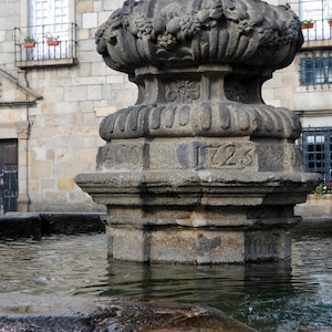 May include: A stone fountain with a carved top and the year 1723 inscribed on the side. The fountain is surrounded by a stone basin filled with water.