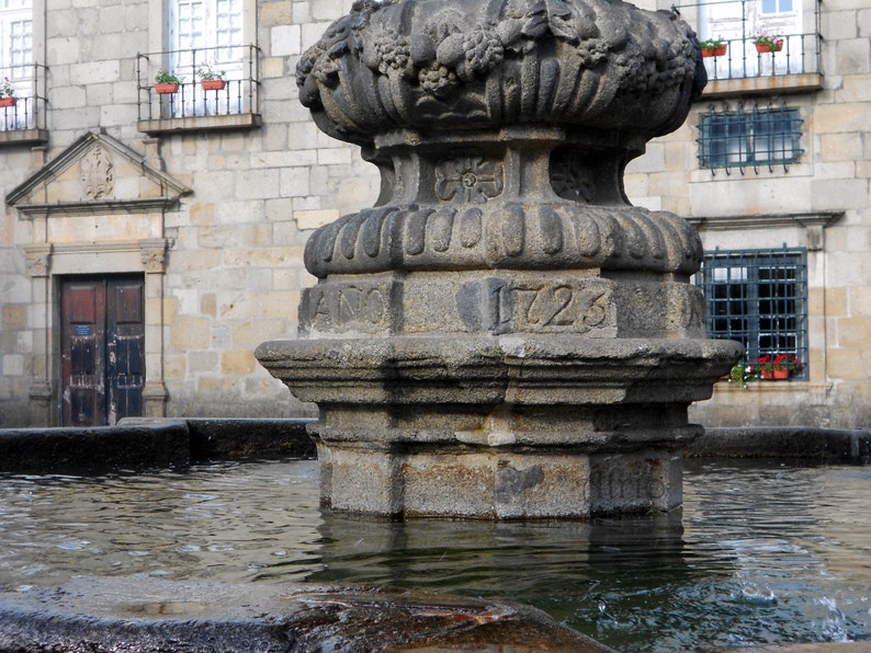 May include: A stone fountain with a carved top and the year 1723 inscribed on the side. The fountain is surrounded by a stone basin filled with water.