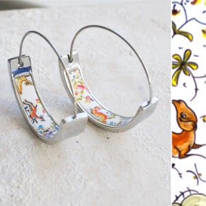 Könnte beinhalten: Ein Paar silberne Creolen mit einem weißen Porzellaneinsatz, der mit einem bunten Blumen- und Tiermotiv verziert ist.