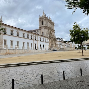 May include: A large, white, stone building with a courtyard in front. The building has a tall, pointed roof and many windows. The courtyard is paved with cobblestones and has a grassy area in the center.