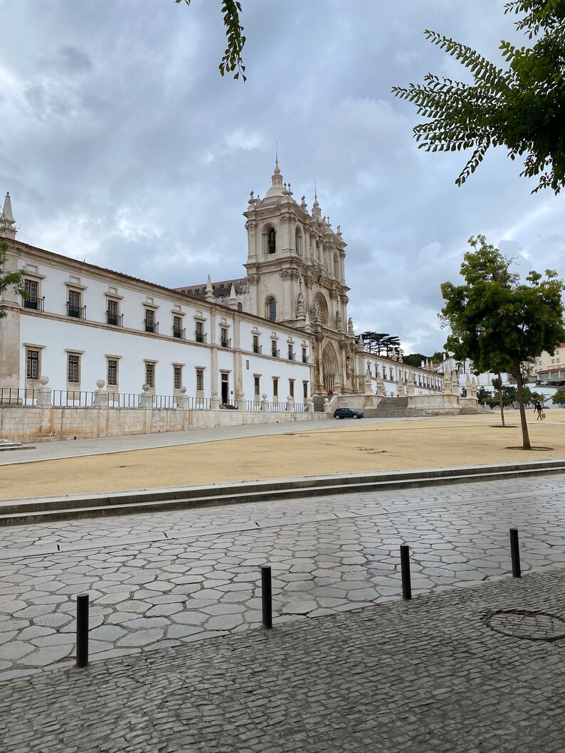May include: A large, white, stone building with a courtyard in front. The building has a tall, pointed roof and many windows. The courtyard is paved with cobblestones and has a grassy area in the center.