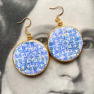 May include: A pair of round earrings with a gold-tone frame. The earrings feature a blue and white patterned design. The design is reminiscent of traditional tile work. The earrings have a hook closure.
