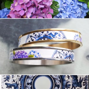 May include: Two bangle bracelets with a white background and blue and gold designs. The top bracelet is gold with a blue and white floral design. The bottom bracelet is silver with a blue and white floral design.