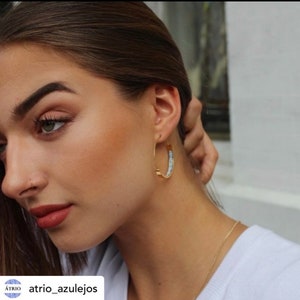 Peut inclure: Gros plan sur le visage d'une femme avec une boucle d'oreille en or &agrave; l'oreille droite. La boucle d'oreille est orn&eacute;e d'un motif blanc et bleu. La femme porte une chemise blanche et a les cheveux tir&eacute;s en arri&egrave;re.