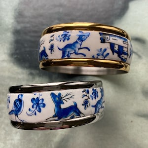 Könnte beinhalten: Zwei dekorative Serviettenringe mit blau-weißem Porzellandesign. Der obere Ring hat eine goldene Blende und zeigt ein blaues Reh und ein blaues Haus. Der untere Ring hat eine silberne Blende und zeigt ein blaues Reh, einen blauen Vogel und blaue Blumen.