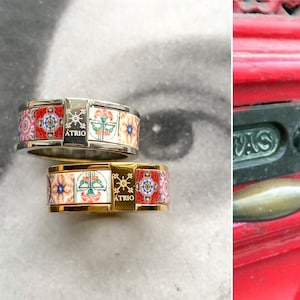 Könnte beinhalten: Zwei Ringe mit einem bunten Fliesenmuster und dem Wort "ATRIO" an der Seite. Der obere Ring ist silber und der untere Ring ist golden.