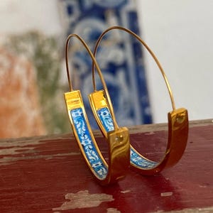 May include: A pair of gold hoop earrings featuring a thin wire post and a curved band inlay with a blue and white floral pattern. The earrings have a polished metallic finish and a circular shape, showcasing a detailed decorative design on the inner surface.