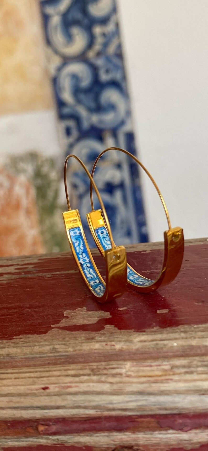 May include: A pair of gold hoop earrings featuring a thin wire post and a curved band inlay with a blue and white floral pattern. The earrings have a polished metallic finish and a circular shape, showcasing a detailed decorative design on the inner surface.