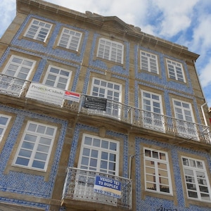 May include: A building with blue and white tile facade. The building has multiple windows and a balcony. A sign on the building reads "VENDO T3 T3 DUPLEX 910 795 356".