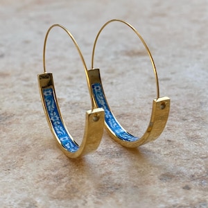 May include: A pair of gold hoop earrings featuring a thin wire post and a curved, flat bottom section. The interior of the bottom section is decorated with a blue and white floral pattern. The earrings have a polished finish and a circular shape.