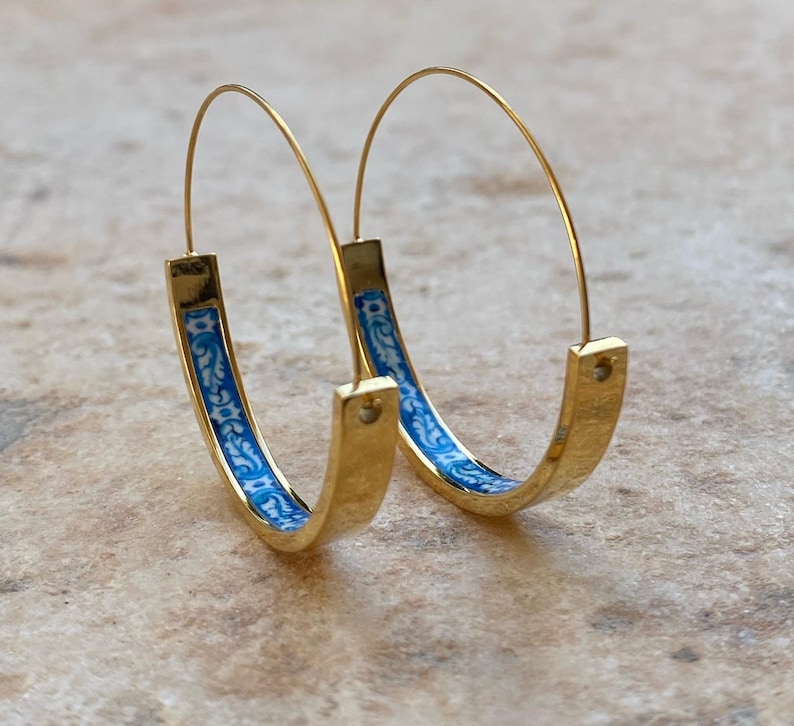 May include: A pair of gold hoop earrings featuring a thin wire post and a curved, flat bottom section. The interior of the bottom section is decorated with a blue and white floral pattern. The earrings have a polished finish and a circular shape.